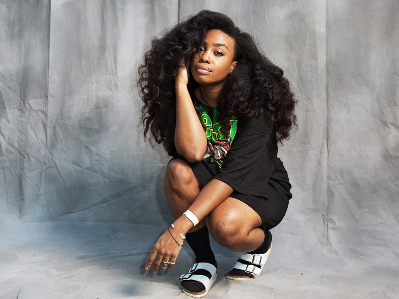SZA poses cooly in a long tee shirt and socks and sandals, crouching low on a wrinkled backdrop © Sniffers 