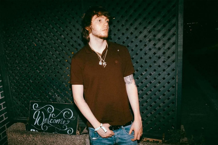 Murda Beatz  has his hands in his pockets looking away with a welcome sign in the background. © XXL